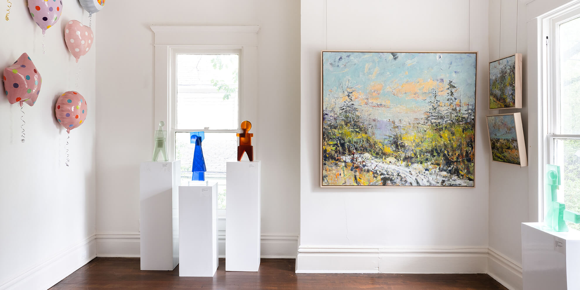 the picture shows a room with white walls, with several pieces of art on each wall. the left wall has ceramic pink balloons covered in multicolored polka dots. the center wall has three white stands holding 3 glass figurines; a light green glass man, and cobalt blue glass woman, and a topaz glass child. next to these three standing figurines is a large landscape canvas painting, with choppy brushstrokes. This piece has a light blue and orange sky, with green and grey trees below and a white water stream at the bottom. the right wall has two small pieces with light blue skies and green forests. next to these pieces, in the foreground, is another glass figurine. it is a mint green woman kicking her leg.
