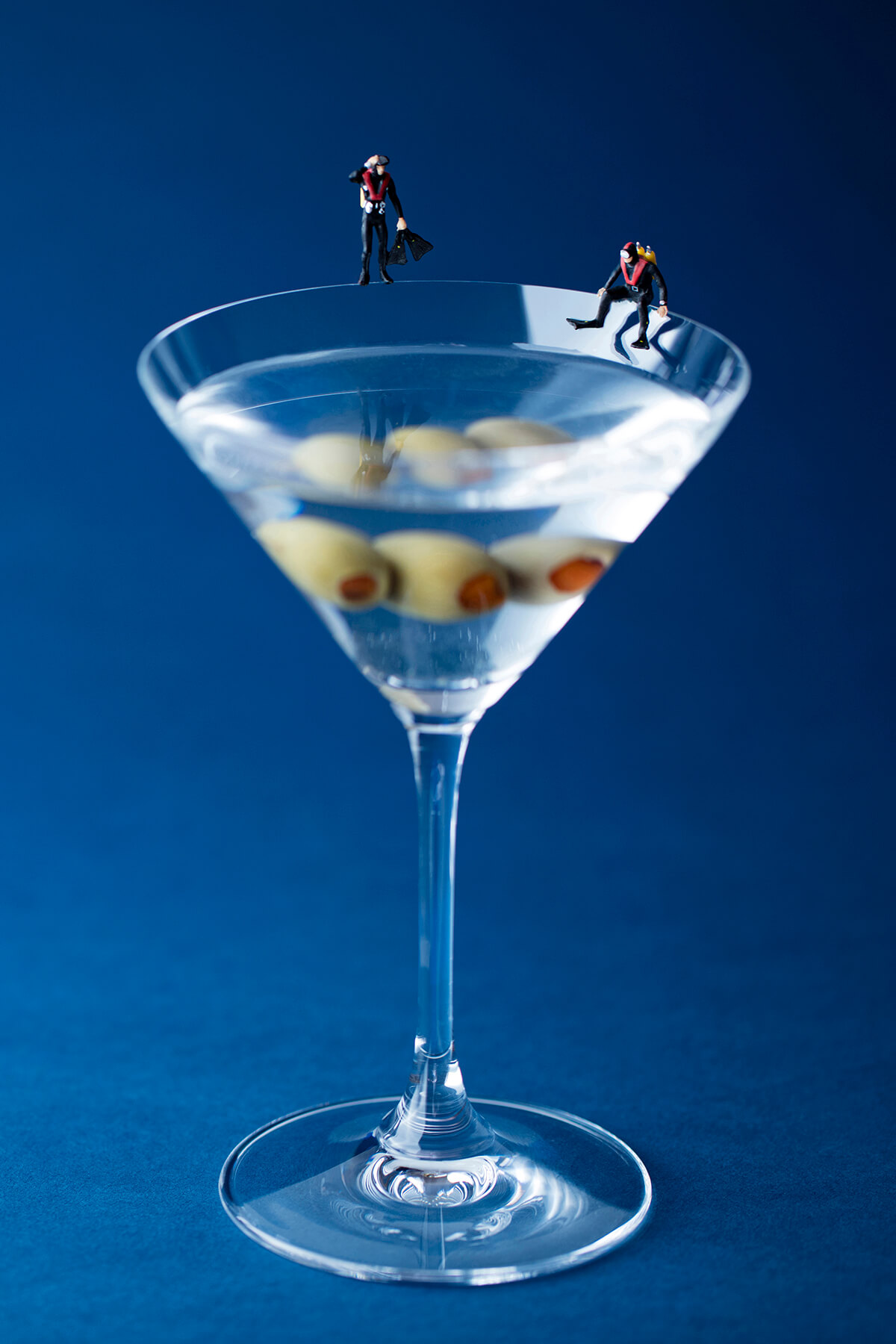 On the rim of a classic crystal martini glass, filled with clear liquid and three pimento-stuffed olives, are two figures, one standing, and the other sitting but both look into the glass dressed out in full wetsuits. The glass is situated in front of a royal blue backdrop.