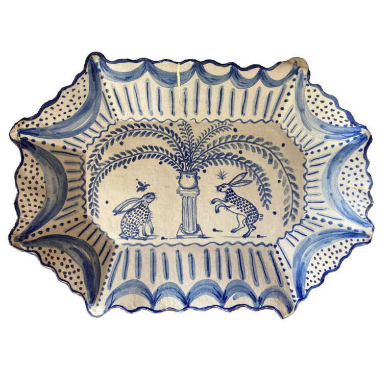 ceramic octagonal platter, cream colored with royal blue detailing. there are swoops and swirls around the border of the platter, with lines and dots decorating it. in the center at the base there is a small scene of two spotted rabbits dancing around a small pillar with a vase on top. the vase has large plume-like plants coming from it.