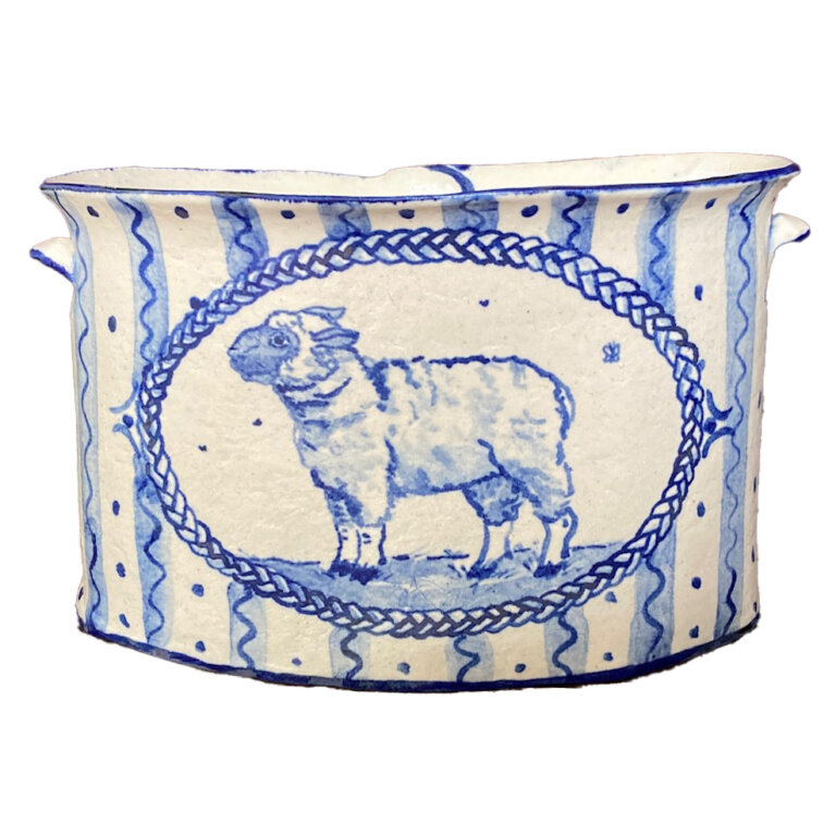 ceramic oval-shaped bowl, cream colored with royal blue detailing. all around the bowl are pale blue vertical stripes with thin darker blue wavy stripes on top. in between each stripe are a few dark blue dots spaced vertically in a line. in the center of the bowl is a thin painted braided frame with a small sheep inside it. the sheep is standing on pale blue grass facing profile to the left. there are two small handles on either side of the bowl.