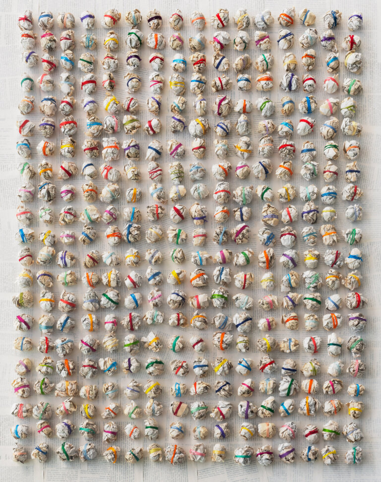 an abstract sculptural piece made up of 336 paper balls arranged in 21 horizontal rows of 16. each ball is wrapped a few times with colorful string in shades of clementine orange, lemon yellow, cherry red, maroon, lime green, sky blue, cobalt blue, lilac, bubblegum pink, magenta, and goldenrod. the background is made up of pages from books covered in a thin layer of white paint