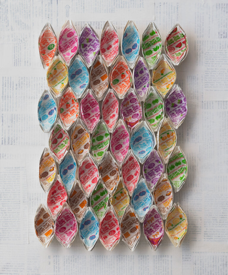 an abstract sculptural art piece with 46 ovular shapes arranged in 7 horizontal rows of 6 or 7. the shapes are made of white paper and lined with different colorful dum dum lollipop wrappers. the background is made up of pages from books covered in a thin layer of white paint