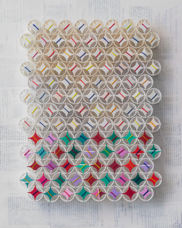 an abstract sculptural art piece with 110 small white paper cups arranged in 13 horizontal rows of 8 or 9, alternating. the top 8 rows of cups each have a white four pointed star on top wrapped in colorful string in shaded of red, lemon yellow, cobalt, coral, orange, and gold. the bottom 5 rows have dark red, cherry red, teal, aqua, lavender, pale pink, magenta, or lilac four pointed stars on top each wrapped with string as well. the background is made up of pages from books covered in a thin layer of white paint