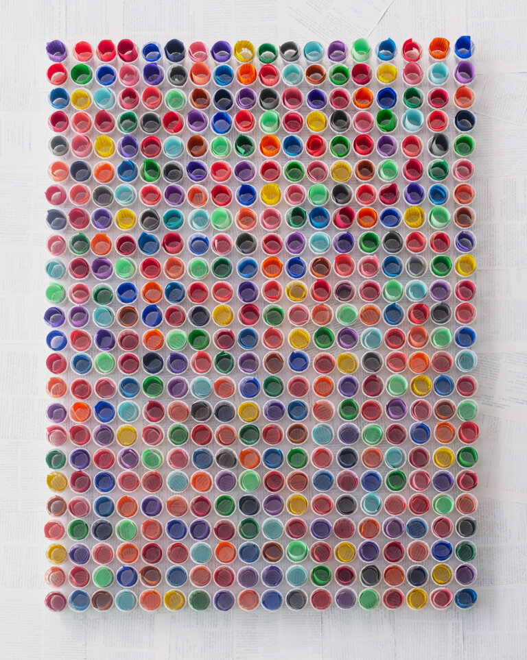 a straight on view of a 3d sculpture piece of small white paper cups arranged in rows, each holding a small roll of colored paper in shades of orange, cobalt, lemon yellow, kelly green, mint, pale aqua, cherry red, slate, mauve, lavender, peach, bubblegum pink, terra cotta, and ultramarine blue