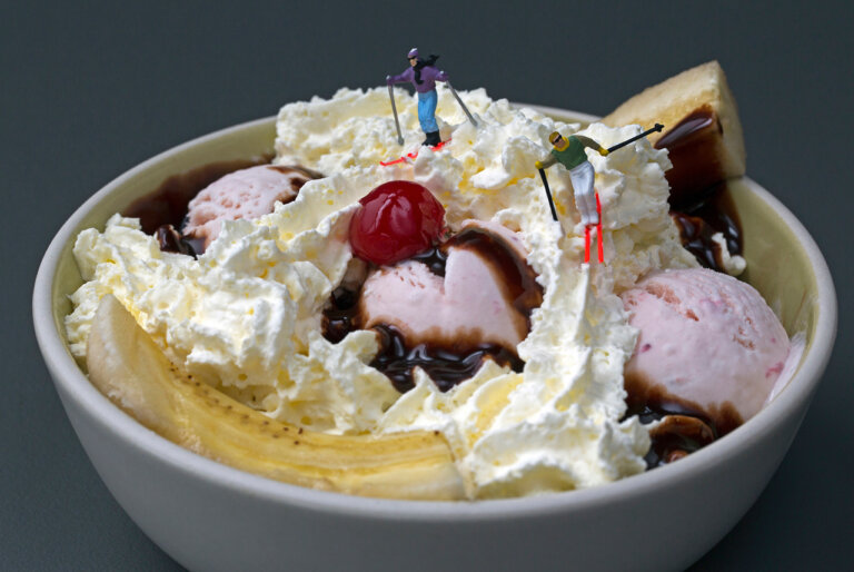 an imaginative photograph of two small skier figurines posed on the whipped cream of a strawberry ice cream banana split as if they are skiing on the whipped cream. the sundae sits in a white bowl with a yellow interior. there is a bright red maraschino cherry in the center and chocolate syrup drizzled around.