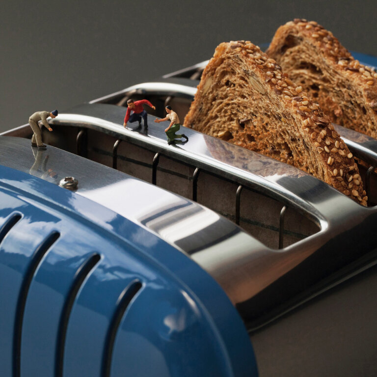 an imaginative photo of a blue and chrome toaster with three toast slots. the back two slots have toast popped up as if it has finished toasting, but the closest slot does not. there are three tiny figures peering over the edge of the empty slot as if they are searching in the "ravine" for the lost piece of toast