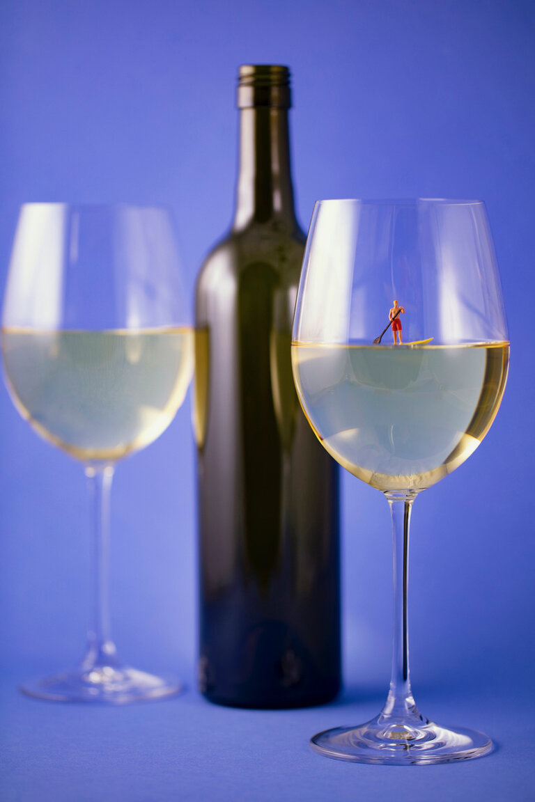 an imaginative photo of two long-stemmed glasses of viognier wine. between the two glasses is a dark green unlabelled wine bottle. on the wine in the glass on the right floats a tiny paddleboarder figurine on a yellow paddle board.