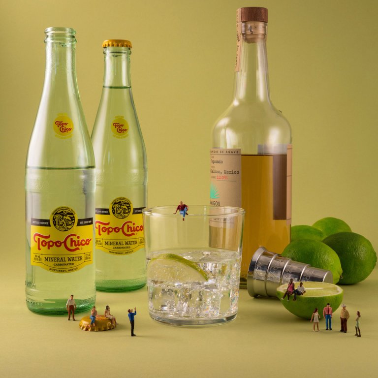 an imaginative photograph featuring two clear glass topo chico sparkling mineral water bottles next to a casamigos reposado bottle that is a little more than halfway full. next to the casamigos bottle are four whole limes and one half lime as well as a silver measuring cup. in the center foreground of the piece is a low ball tumbler that is half full of ice, and clear liquid, as well as a small lime wedge. there are tiny people posed around the scene which add a whimsical air to this photograph