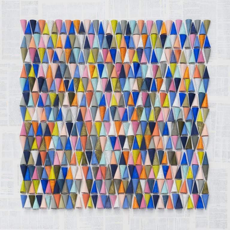 an abstract geometric piece consisting of 13 rows of paper cones lying on their sides arranged in neat rows of 41. the cones are painted in shades of cobalt, powder blue, mint, tangerine, citron, bubblegum pink, white, olive, navy, and gray. there is a background of washed out book pages behind the square of cone rows.
