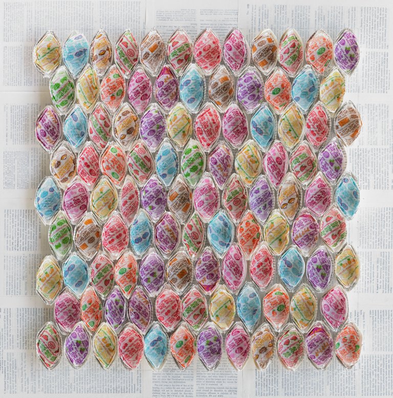 an abstract sculptural art piece with 46 ovular shapes arranged in 9 horizontal rows of 12 or 11. the shapes are made of white paper and lined with different colorful dum dum lollipop wrappers. the background is made up of pages from books covered in a thin layer of white paint