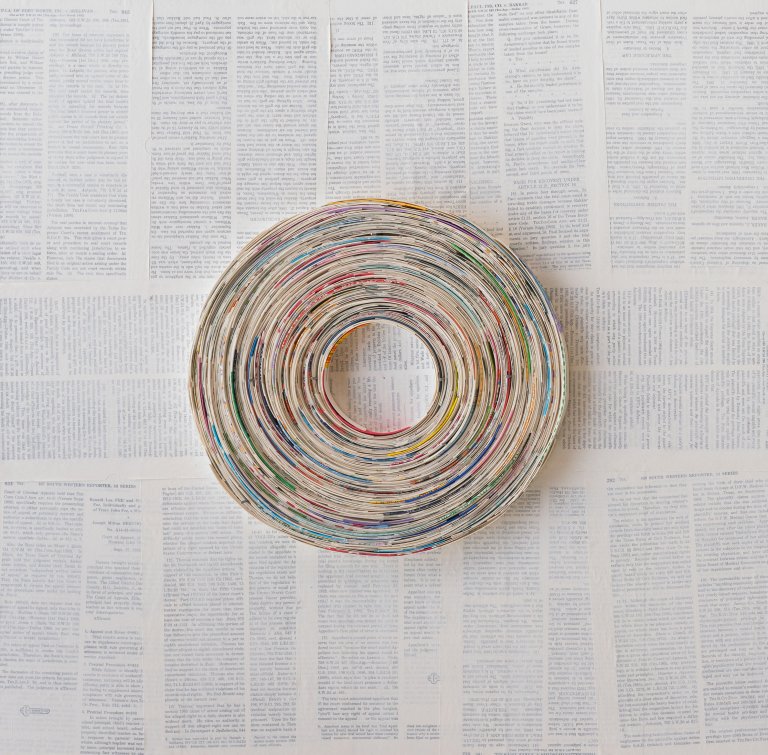 an abstract collaged piece that resembles a vinyl record. there are small pieces of paper swirled in a circle shape. the background is collaged with book pages and has a thin layer of white paint over top that creates a faded look