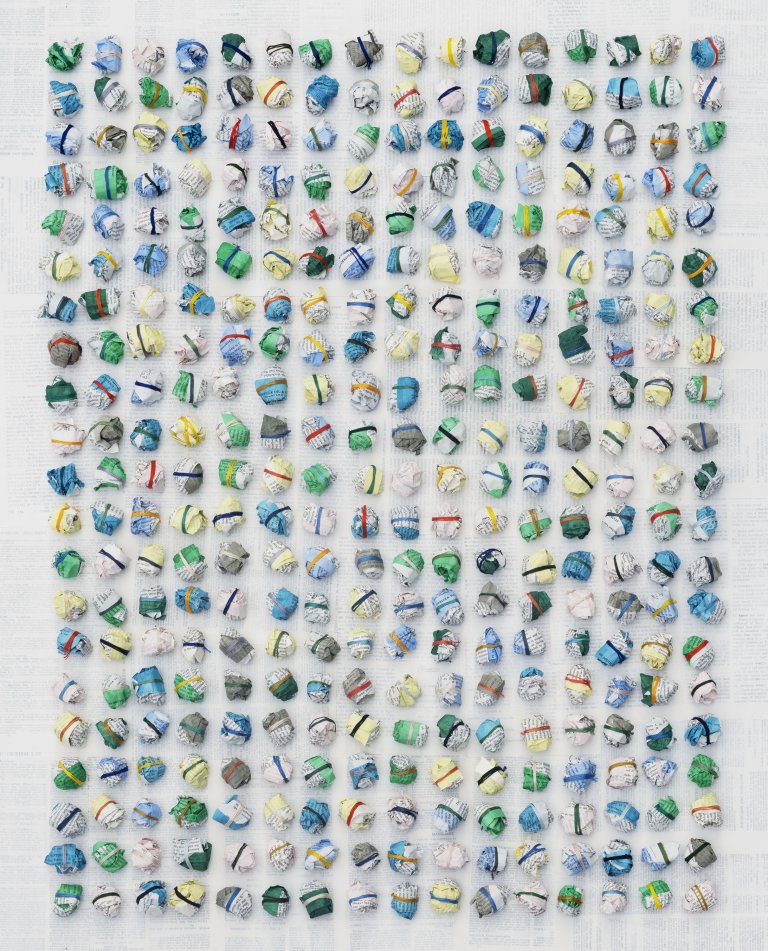 an abstract sculptural piece made up of 336 paper balls arranged in 21 horizontal rows of 16. each ball is wrapped a few times with colorful string in shades of lime green, lemon yellow, emerald green, kelly green, pale yellow, sky blue, cobalt blue, lilac, bubblegum pink, magenta, and soft gray. the background is made up of pages from books covered in a thin layer of white paint