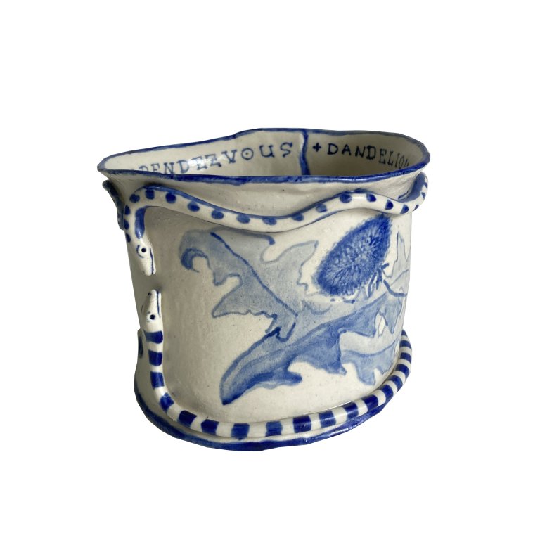a ceramic bowl that is cream colored with cobalt blue detailing. there is a beautiful dandelion leaf and blossom pattern on one side. on the outside of the bow there are two snakes that wrap around the bowl. one snake is spotted, and the other is striped. the top and bottom rims of the bowl are lined with blue. the inside of the bowl bears the words "DANDELION RENDEZVOUS" in all capital letters.