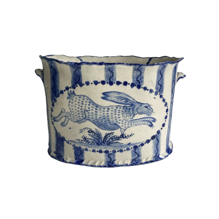 a ceramic bowl that is cream colored with cobalt blue detailing. the bowl has a horizontal oval painted on the front. inside is a leaping hare that is spotted and viewed in profile leaping over a small leafy plant. the background behind the oval is pale blue vertical stripes with a dark blue wavy line inside. there are two small tabs on the bowl, one on each side, that look like small handles. the top rim of the bowl is lined with blue.