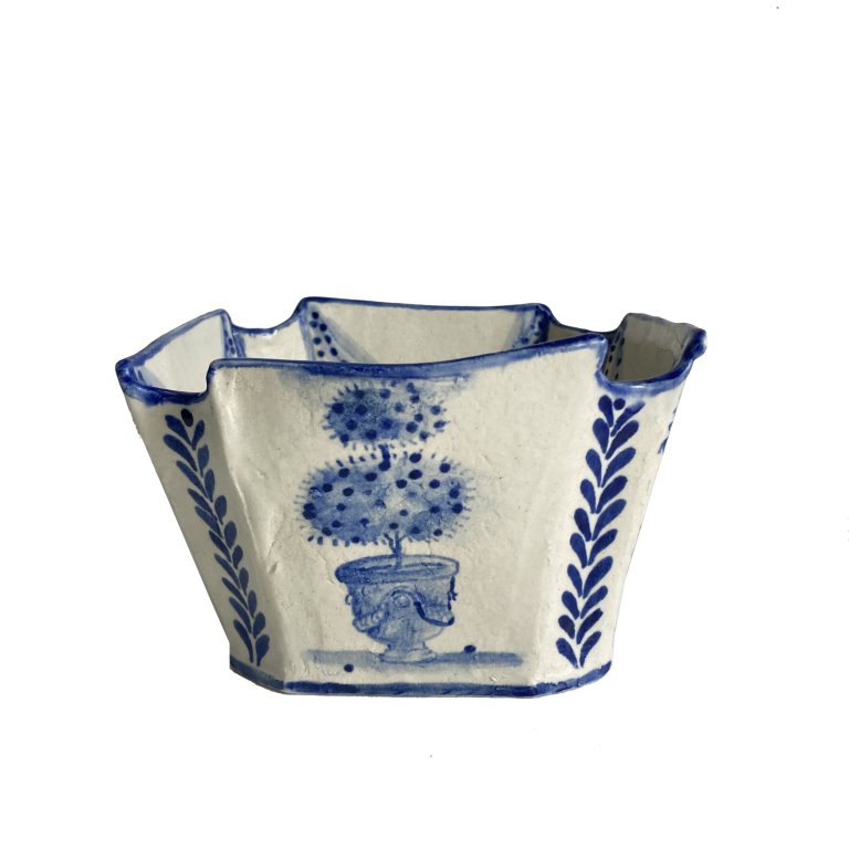 a ceramic bowl that is cream colored with cobalt blue detailing. the rim of the bowl has four divots, one in each corner, and it's rimmed in blue. there is a two-tiered topiary on the front in an ornate pot. the topiary is a fruit-bearing plant, and has small round fruits. there are two fruits on the ground next to the planter. there is a leaf-like vertical pattern on either side of the topiary.