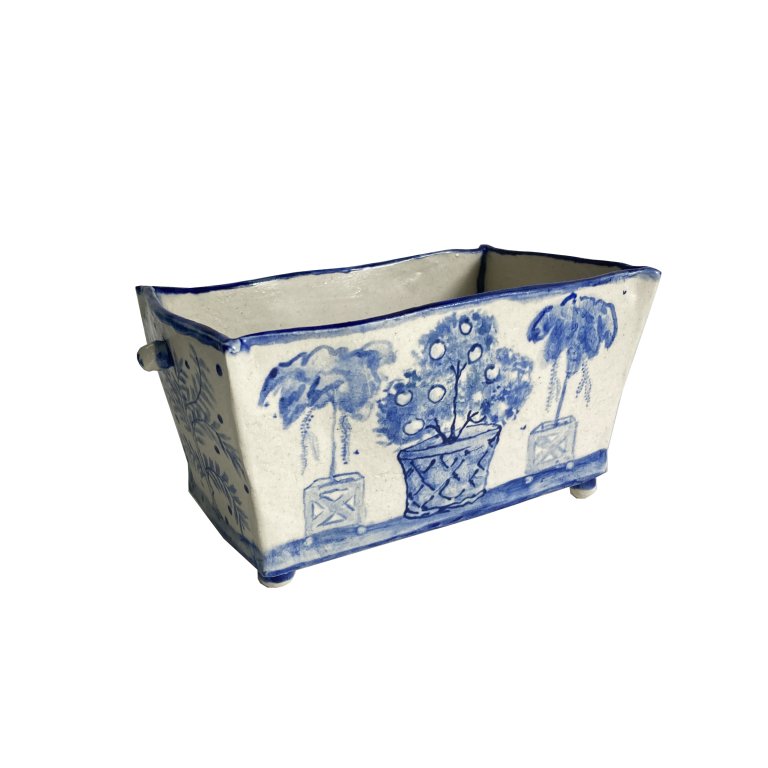 a ceramic bowl that is cream colored with cobalt blue detailing. the bowl is rectangular. on the long side there are three planters with trees inside. the left and right planters are similar, with a crossed "x" design and a tree with a thin trunk and fluffy foliage with small vines hanging down. the middle planter has a fruit-bearing tree which is lower and rounder. the short side of the bowl has a leafy vine detail. the bottom and top edges of the bowl are rimmed in blue. there are small spheres under the four bottom corners of the bowl, creating small legs to support the bowl. there is a small tab that looks like a little handle on each of the short sides near the top.