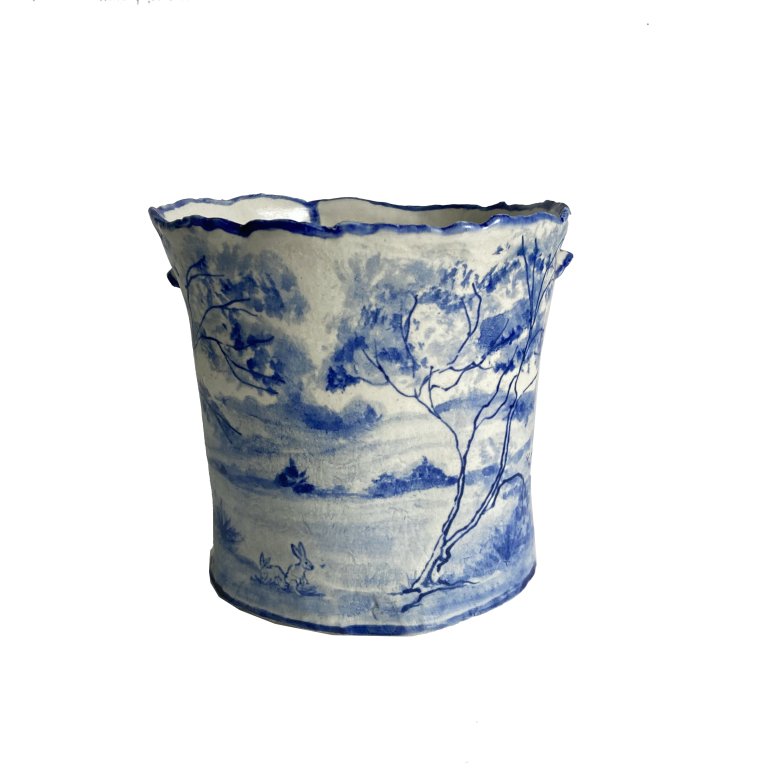 a ceramic bowl that is cream colored with cobalt blue detailing. there is a beautiful landscape painted on the side of the bowl, depicting a large spindly tree with a thin trunk and soft fluffy foliage. there are small shrubs on the ground, and two small rabbits. the top edge of the bowl is lined with blue.