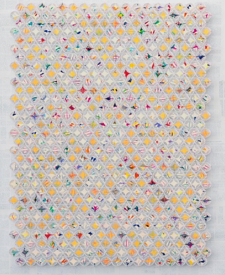 an abstract art piece that is constructed of small paper sample cups arranged in horizontal rows with small four-pointed stars cut out of paper resting on top of each cup. there is an overwhelming amount of yellow stars, but these are punctuated with multicolored stars as well.