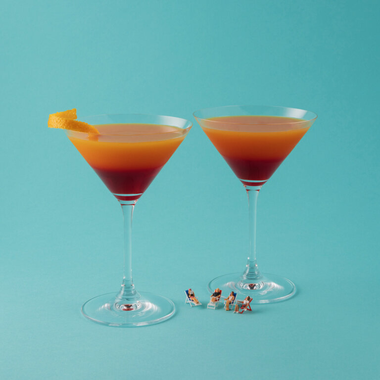 an artistic photograph with a turquoise background showing two cocktails in clear glass martini glasses. the cocktails are on ombre of deep red to light orange. the cocktail on the left has a twist of orange peel. at the base of the cocktails are four miniature figures in lawn chairs, reading and arranged so that it looks like they are casually chatting about books.