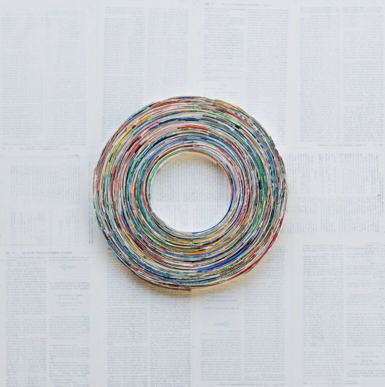 an abstract collaged piece that resembles a vinyl record. there are small pieces of paper swirled in a circle shape. the background is collaged with book pages and has a thin layer of white paint over top that creates a faded look