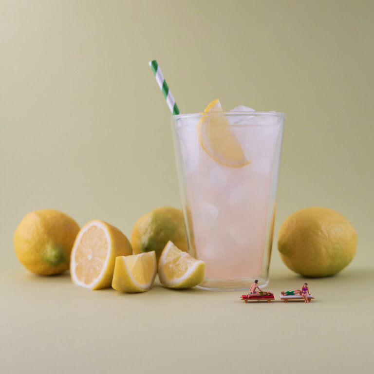 an artistic photograph styled by artist christopher boffoli showing a clear glass of iced pink lemonade with a green and white striped straw and a single lemon slice. the glass rests on a muted pale yellow background. there are three whole lemons resting next to the glass, and a half lemon and two wedges resting next to these. there are four figures in swim attire on two chaise lounges in front of the glass