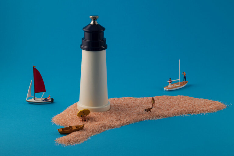 a playful artistic photograph of a salt grinder and pink salt arranged against a blue background to look like a small beach and a light house. there is a small rowboat docked on the salt "beach" with two small figures under a yellow umbrella next to it. further up the "beach" there is a figure in yellow walking with a brown and white dog. in the "water" around the sand there are two sailboats: one with the sail raised and a figure riding leisurely inside, and another with two figures working to raise the sail.