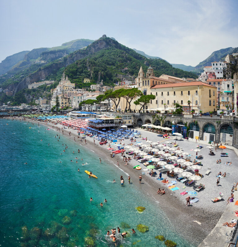 a photograph of the amalfi coast showing turquoise water and a gray pebbled beach with rows of umbrellas and beach goers. behind the beach is the town of amalfi and luscious green mountains