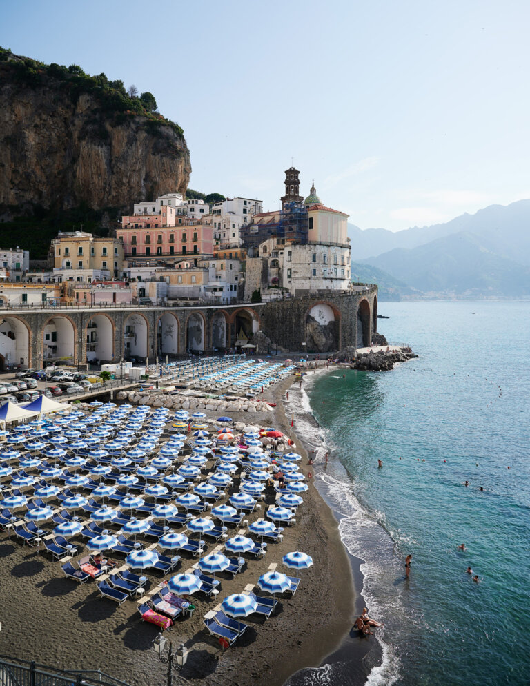 a photograph of the italian coast showing turquoise water and a gray pebbled beach with rows of umbrellas and beach goers. behind the beach is the town of atrani and luscious green mountains