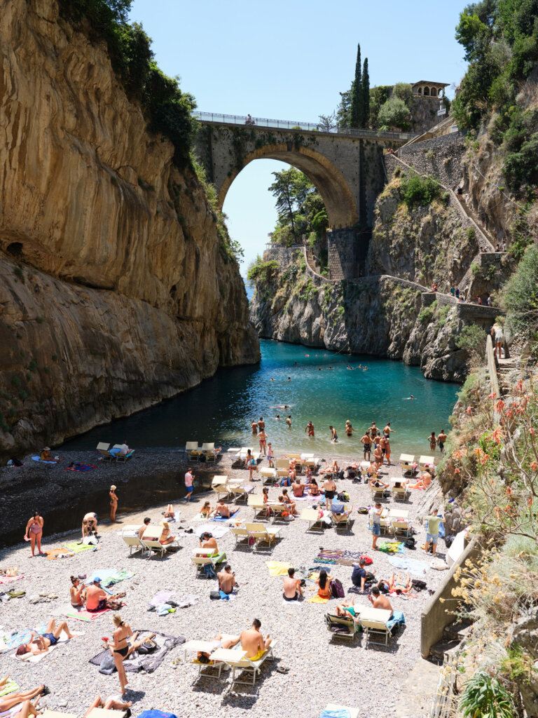 a photograph of a rock pebble beach and turquoise water between two cliff faces. the beach is scattered with beachgoers on their towels. there is a bridge in the distance connecting the two cliffs with an arch. the sky is blue above and very sunny.
