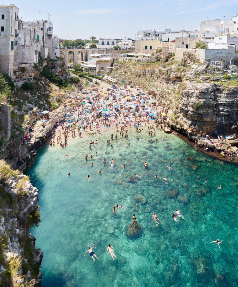 a photograph of teal water and rocks by the beach in puglia. there are people on the beach and in the water. in the distance there is the town of puglia