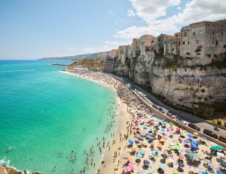 a photograph showing a view of the coast in tropea. there is bright teal water washing up on a sandy beach. the town is next to the beach, with a steep cliff face that leads up to buildings. there is a narrow highway lined with cars separating the cliff face from the beach. the beach is covered in colorful umbrellas. there are beachgoers scattered on the beach and in the water