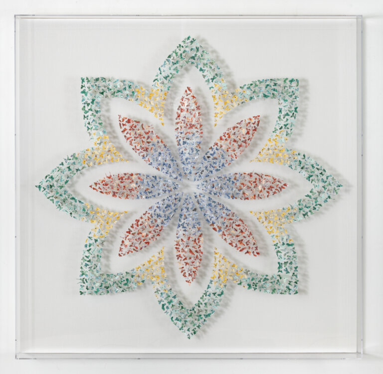a 3d art piece depicting a floral shape made of tiny butterflies cut out and mounted on pins. the outer floral shape is in green and yellow, and the inner flower has a blue center with red-tips.