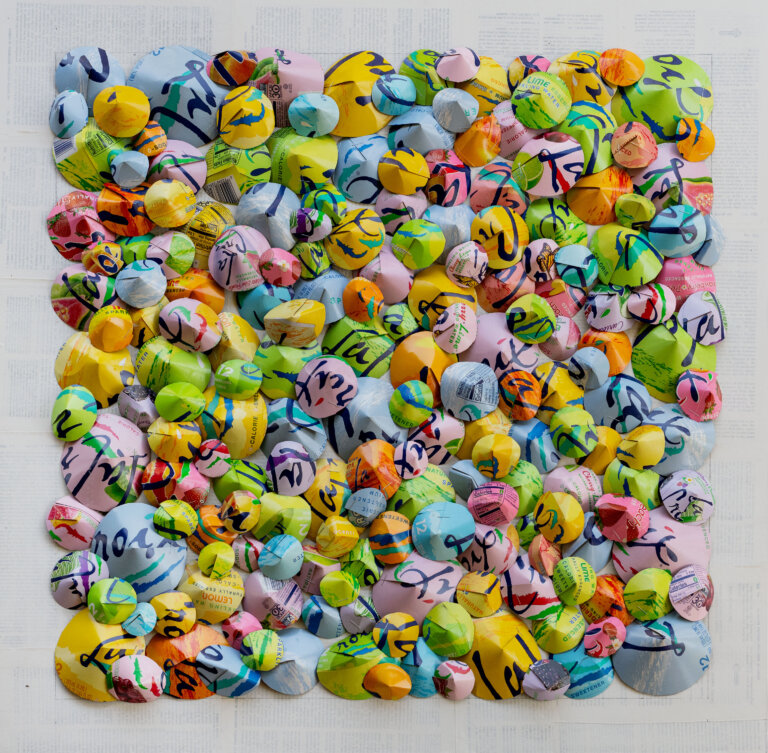 a collage made of wide flat cones of la croix box cardboard in all different colors that overlap and layer with each other to create a square shape. the background is made up of pages from books covered in a thin layer of white paint