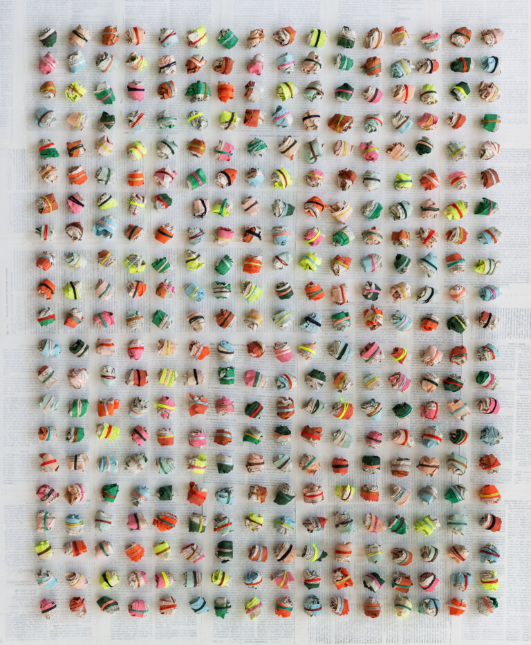 an abstract sculptural piece made up of paper balls arranged in horizontal rows. each ball is wrapped a few times with colorful string in shades of cherry red, lemon yellow, emerald green, vermillion, burgundy, sky blue, cobalt blue, and magenta. the background is made up of pages from books covered in a thin layer of white paint