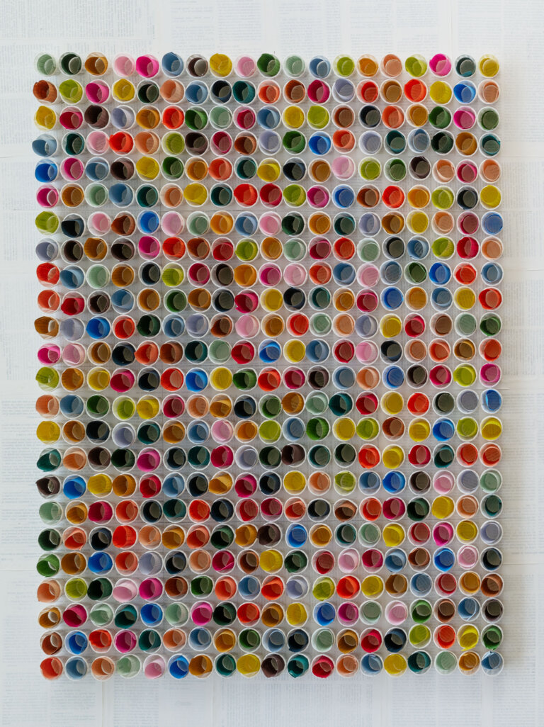 a straight on view of a 3d sculpture piece of small white paper cups arranged in rows, each holding a small roll of colored paper in shades of orange, cobalt, lemon yellow, kelly green, mint, pale aqua, cherry red, slate, mauve, lavender, peach, bubblegum pink, terra cotta, and ultramarine blue