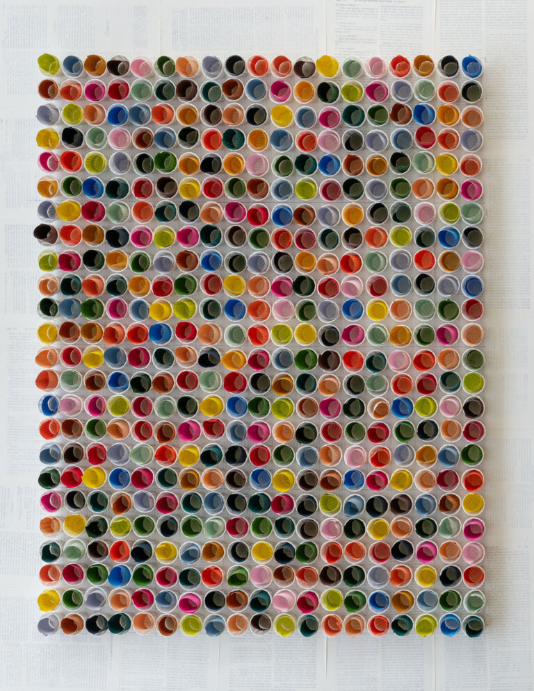a straight on view of a 3d sculpture piece of small white paper cups arranged in rows, each holding a small roll of colored paper in shades of orange, cobalt, lemon yellow, kelly green, mint, pale aqua, cherry red, slate, mauve, lavender, peach, bubblegum pink, terra cotta, and ultramarine blue