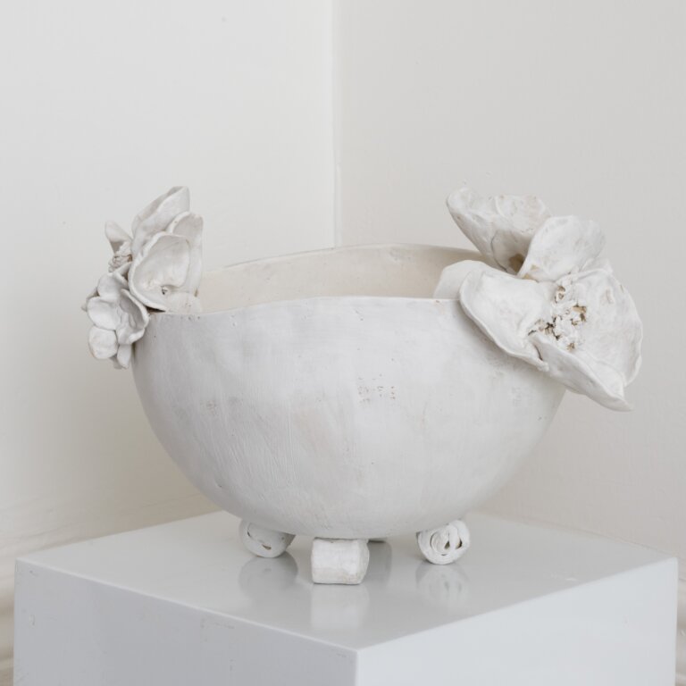 a hand-built stoneware vessel with a rounded, bowl-like form and a gently irregular rim. the surface is finished in a matte, chalky white, with subtle variations and faint marks that emphasize its handmade character. sculpted floral elements sit on opposite sides of the rim, their soft, layered petals adding dimensional contrast and a delicate, organic touch. the piece is elevated on small, rolled feet, reinforcing its sculptural presence. overall, it feels quiet, refined, and tactile, with a balance of simplicity and expressive detail.
