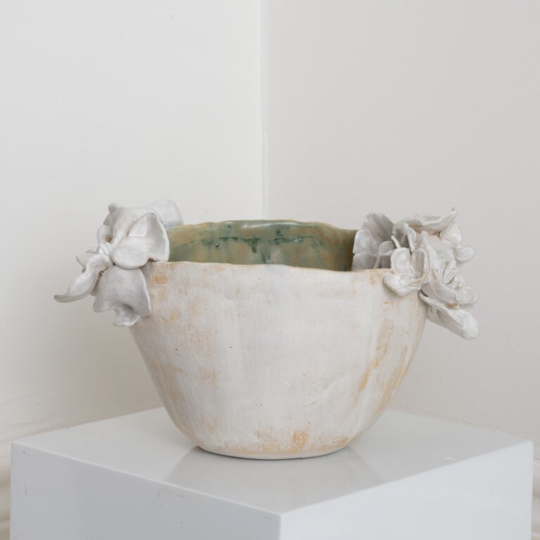 a hand-built stoneware vessel with a softly rounded, tapering form and a gently irregular rim. the exterior is finished in a matte, chalky white with warm clay undertones peeking through, giving it a subtly weathered, tactile surface. sculpted floral elements are applied at opposite sides of the rim, their layered petals adding dimensional contrast to the smooth body. Inside, a soft green glaze pools and drips slightly, introducing a quiet color accent. the piece feels organic and balanced, blending sculptural detail with a restrained, earthy elegance.